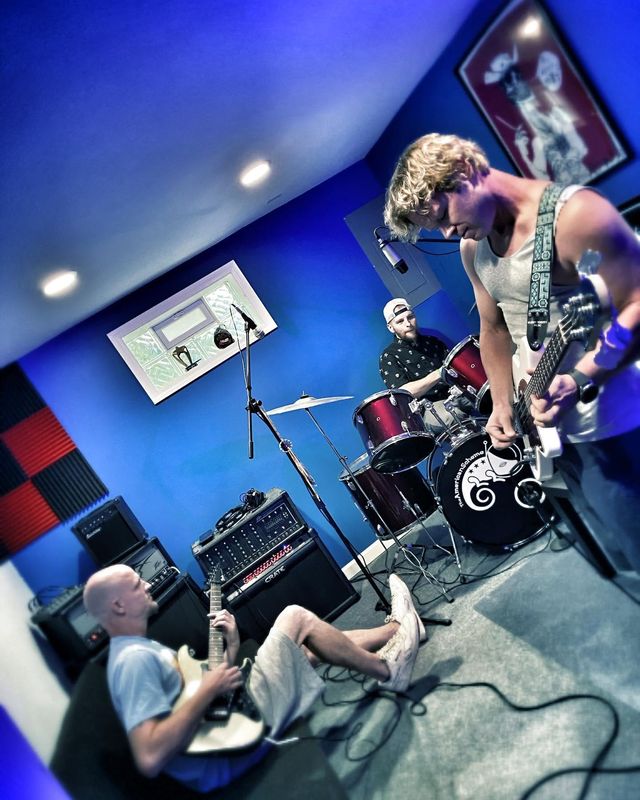 The band playing guitar, bass and drums in a blue practice room